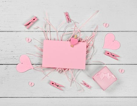 Close up one blank pink Valentine paper card note with heart shaped wooden pin and gift box over white wooden table background, flat lay, elevated top view, directly above