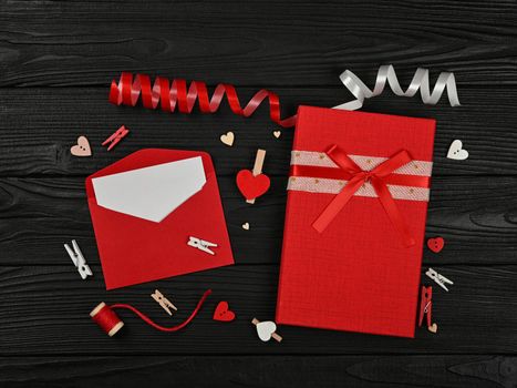 Preparing Valentine gifts, red boxes, hearts, twine, clothespins and note in paper envelope on black wooden table background, close up flat lay, elevated top view, directly above