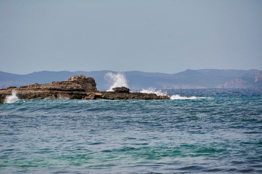 Rocks on the shore hit by the waves. Balearic Islands, Mediterranean Sea, blue sky, splashes of water.