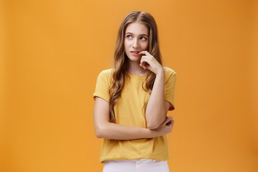 Dreamy and focused attractive young common slender girl with wavy beautiful hair touching lip gazing at upper left corner thoughtful, thinking spacing out while daydreaming over orange wall. Body language concept