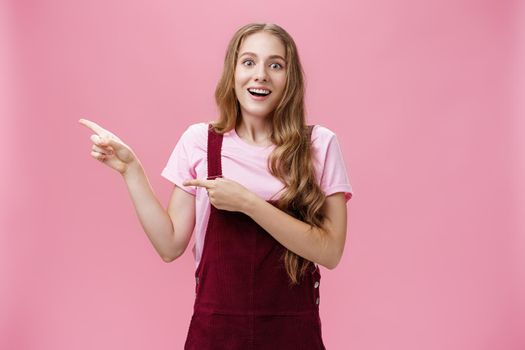 Lifestyle. Woman looking with impressed and amazed look showing cool think on left side of copy space smiling fascinated looking with admiration at camera standing delighted and surprised over pink background.