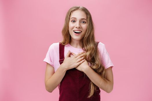 Portrait of thankful and pleased happy young girl receiving surprise gift on b-day pressing palms to hear in gratefyl gesture open mouth from amazement and joy thanking over pink background. Body language and emotions concept