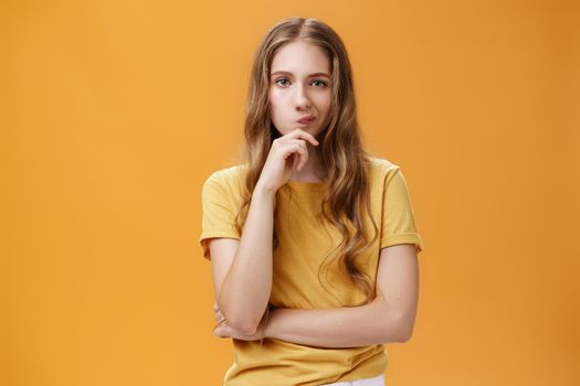 Hmm choices difficult. Portrait of focus serious-looking creative young female with wavy hair smirking holding hand on chin in thoughtful pose, making up decision, thinking over orange background.