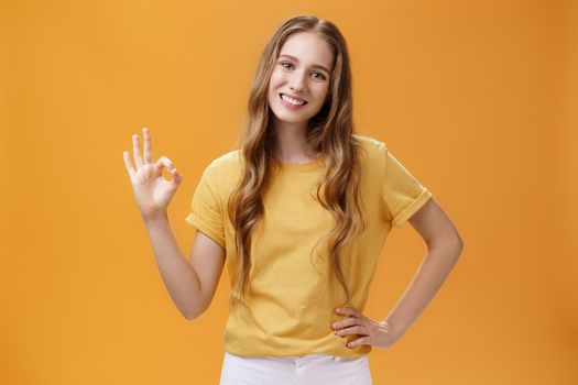 Girl showing positive answer with body language. Charming friendly-looking young female student with natural wavy hairstyle holding hand on hip making ok gesture and smiling over orange wall.