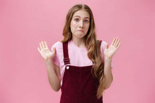 I am not involved. Portrait of unaware and clueless innocent cute woman raising palms in surrender pulling lips down as if being confused and uncertain what happening having no idea over pink wall.