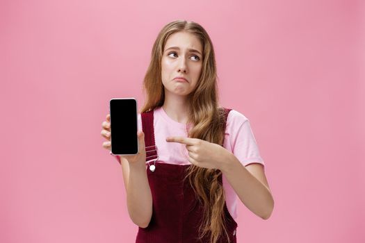 Indoor shot of gloomy displeased and disappointed cute young girl holding smartphone pointing at cellphone screen making upset grimace as if being bothered and dissatisfied against pink wall. Emotions and technology concept