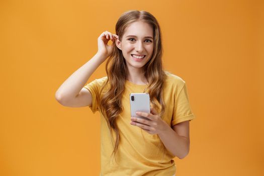 Lifestyle. Waist-up shot of cute and friendly-looking nice young girl with wavy hairstyle putting hair strand behind ear holding smartphone smiling broadly with joyful expression at camera over orange background.