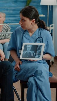 Woman nurse with x ray on tablet showing scan to old patient in nursing home. Medical assistant holding osteopathy radiography on device for disabled man in wheelchair. Person getting diagnosis