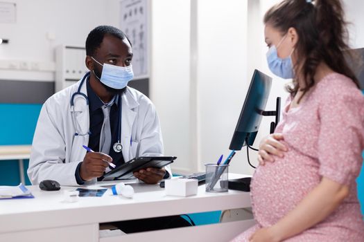 Pregnant adult meeting with doctor at checkup appointment during coronavirus pandemic. Patient with pregnancy talking to practitioner about birth and medical care, wearing face masks.