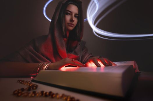 Young caucasian attractive fortune teller witch astrologer woman in a dark hoodie with a hood with luminous palms reads the book of the fate of magic. Glowing circles of spirits in the air. Looking into the camera. Copy space