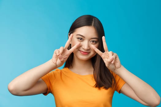 Tender cute asian woman short dark haircut show peace victory signs near cheeks, posing joyfully grinning optimistic, determined win, tilt head coquettish, stand blue background cheerful.