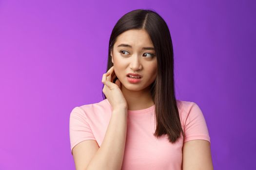 Insecure anxious young timid asian girl feel nervous scared, scratch neck worried, look sideways alarmed, feel danger, standing upset purple background distressed suffer discomfort.