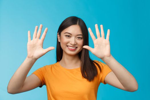Close-up friendly attractive cheerful stylish asian girl raise hands show ten fingers, order dozen, smiling joyfully, judging good job tenth score, stand blue background upbeat. Copy space