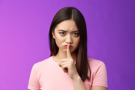 Close-up offended serious-looking upset asian young girl frowning cannot focus disturbed loud conversation, hush stare camera unsatisfied, show shush sign press finger lips, purple background.