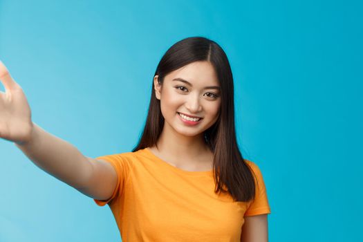 Close-up attractive friendly modern vietnamese girl extend hand, taking selfie, smiling joyfully, make photograph smartphone, hold camera arm, stand blue background joyful video-call friend.