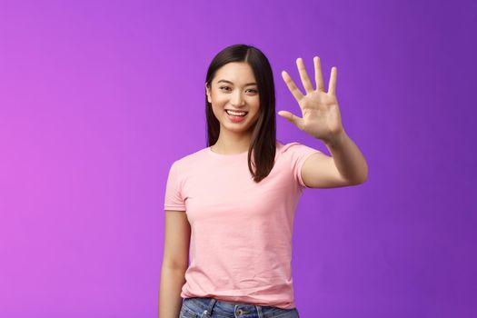 Friendly cute asian tender girl raise palm waving goodbye, smiling joyfully, farewell, gladly welcome friends inviting come in, saying hi hello, greeting, stand purple background. Copy space