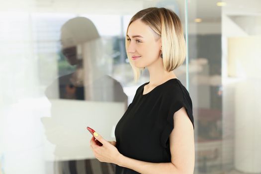 Portrait of attractive young blonde businesswoman in stylish black blouse with smartphone in company office. Business, corporation, success, career concept
