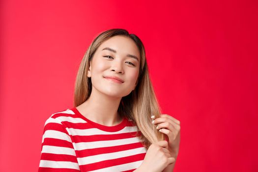 Close-up satisfied cute carefree asian blond girl feeling delighted and pleased after haircare procedure touch healthy hair tilt head smiling broadly like new hairdresser, stand red background.