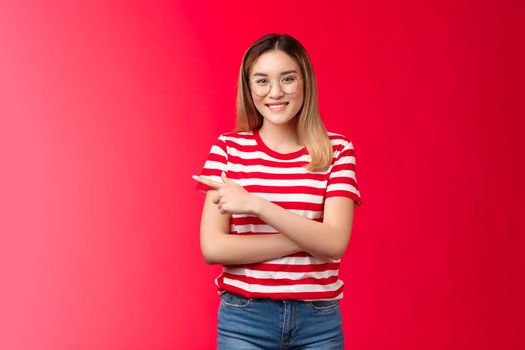 Cheerful confident female asian blond femenist wear glasses pointing left casually discuss advertisement, introduce cool place promo stand red background relaxed, smiling happily camera.
