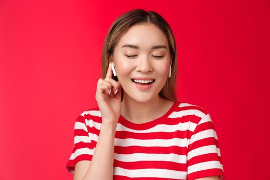 Carefree delighted asian gentle woman close eyes satisfaction smiling broadly wear wireless earphones touch earbud volume up enjoy perfect sound headphones, listen music happily red background.