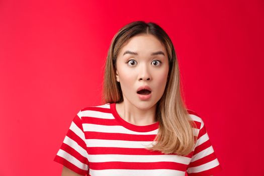 Close-up shocked alarmed speechless asian blond woman drop jaw, gasping impressed, hear shocking stunning rumor, open mouth astonished, react shook news, stand red background. Copy space
