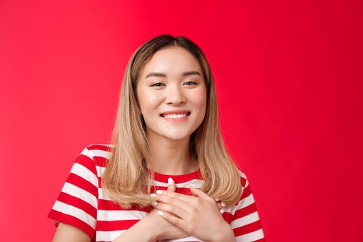 Close-up tender touched young charming asian blond girl press palms heart thankful smiling broadly appreciate nice romantic compliments share heartwarming feelings, stand happy red background.