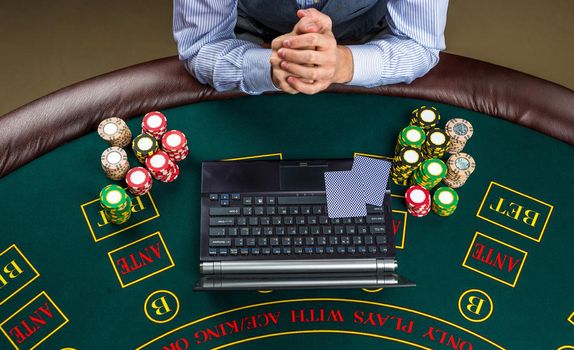 casino, online gambling, technology and people concept - close up of poker player with playing cards, laptop and chips at green casino table