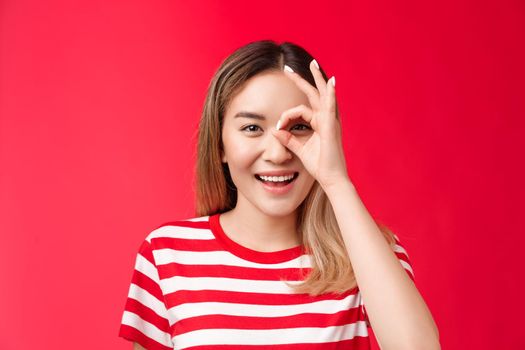Close-up upbeat positive asian girl look through ok sign smiling broadly, enjoy positive vibes, judging, grinning spend great summer holidays, approve cool idea, stand red background.