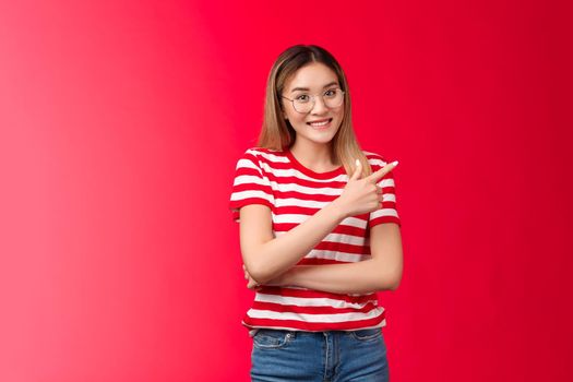 Cheerful friendly asian female blond hairstyle wear striped summer t-shirt glasses, pointing right showing you cool place smiling broadly, stand red background relaxed, introduce promo offer.