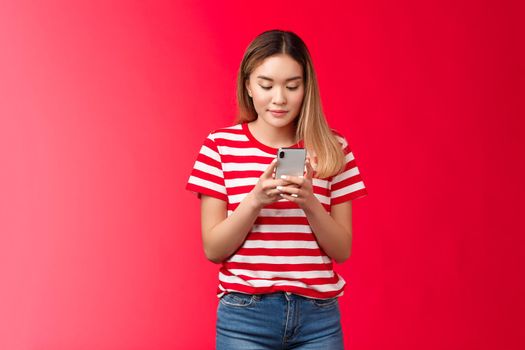 Popular asian female lifestyle blogger posting photo online hold smartphone look phone display scroll feed, woman smiling delighted relaxed using gadget stand red background. Copy space