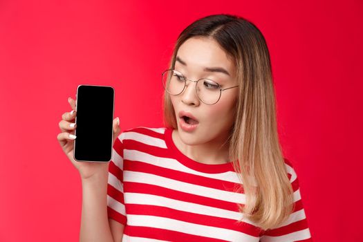 Close-up wondered surprised attractive asian blond girl wear glasses hold smartphone looking impressed amused phone screen, open mouth interesting app telephone display, stand red background.