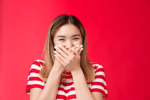 Close-up cute tender silly asian girl giggling, fool around having fun, press palms lips promise not tell secret, trying hold laugh enjoy friendly moment, stand red background grin with eyes.