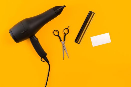 Black hair dryer, comb and scissors on yellow paper background. Top view. Copy space. Still life. Mock-up. Flat lay