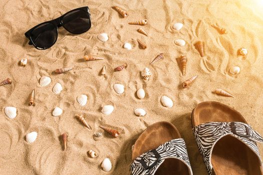 Black flip-flops and sunglass, seashell on sand. With place for your text. Top view. Copy space. Sun flare