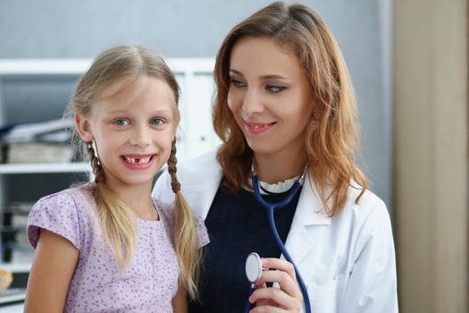 Portrait of smiling professional pediatrician posing with cute little girl on appointment. Doctor hold stethoscope tool for examining. Healthcare concept