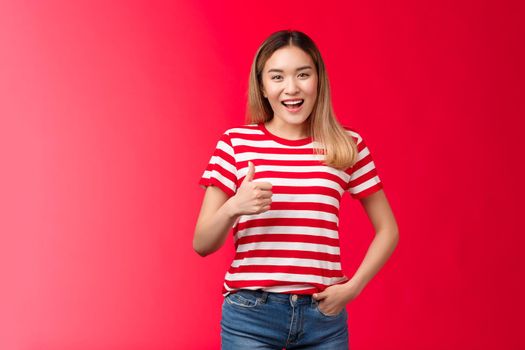 Nice idea my approval. Pleased satisfied good-looking blond asian girl show thumb up and smile, give positive reply, agree wish good luck, judge excellent work, stand red background.