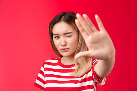 Close-up serious-looking determined asian blond woman pull hand forward show stop taboo sign, concentrating gaze on camera look with one eye, demanding stop, tell enough, say no, red background.
