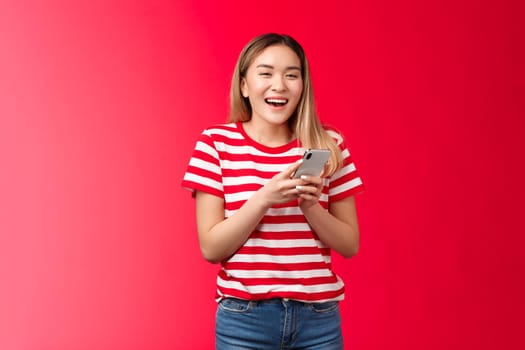 Happy cheerful asian blond girl laughing using smartphone, girl hold device smiling broadly, look camera amused, texting girlfriends, reading funny internet post, stand red background carefree.
