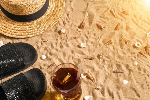 Summer beachwear, flip flops, hat, cold drink in a glass and seashells on sand beach. Top view. Copy space. Sun flare