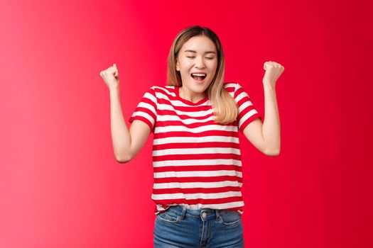 Hooray I did it. Triumphing cute cheerful asian blond girl make fist pumps closed eyes happy broad smile, celebrating accomplishment feel satisfaction finally winning achieve success red background.