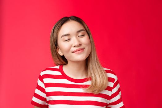 Close-up romantic tender happy asian woman tilt head close eyes joyfully smiling recalling nice lovely moment, daydreaming delicious coffee with dessert, stand red background relaxed cheerful.