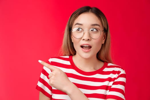 Amused wondered cute asian blond modern girl surprised, looking pointing left intrerested open mouth fascinated amazed, checking out interesting event, attend curious exhibition, red background.