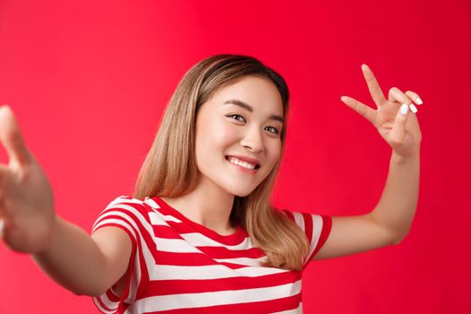 Friendly outgoing cute asian girl blond short hairstyle extend arm, hold smartphone camera, tilt head joyfully show peace victory sign, taking selfie photographing herself, stand red background.