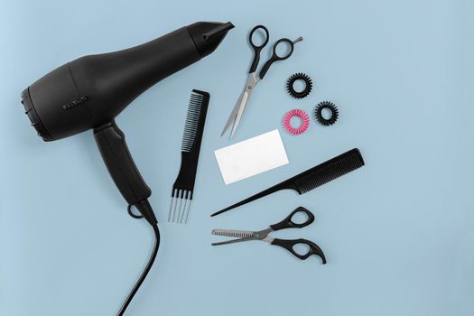 Black hair dryer, comb and scissors on blue paper background. Top view. Copy space. Still life. Mock-up. Flat lay