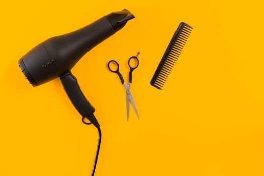 Black hair dryer, comb and scissors on yellow paper background. Top view. Copy space. Still life. Mock-up. Flat lay