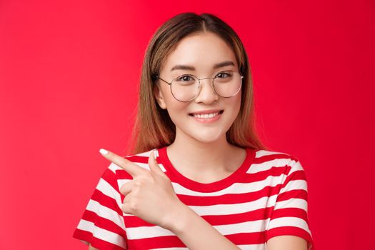 Good opportunity behind corner. Cute asian girl wearing glasses striped summer t-shirt pointing left, smiling toothy friendly expression, woman introduce promo indicating good choice, red background.