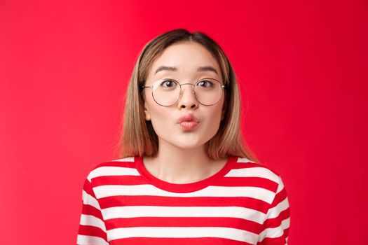 Cute pretty asian blond modern teenage girl folding lips anticipating tender romantic moment kiss, look camera wear glasses, blowing mwah, stand silly red background, feel passionate coquettish.