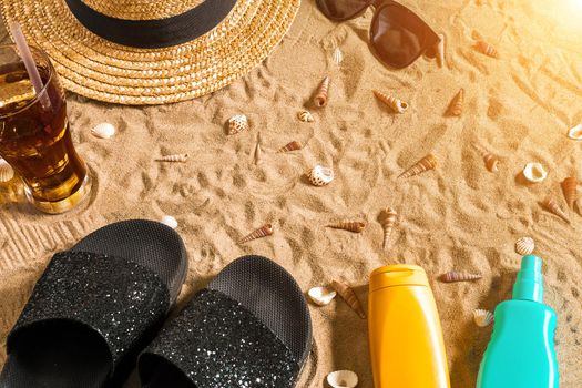 Summer beachwear, flip flops, hat, cold drink in a glass and seashells on sand beach. Top view. Copy space. Sun flare