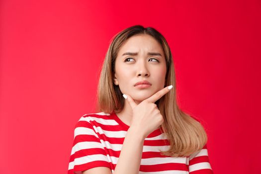 Close-up suspicious thoughtful girlfriend pondering, touch chin squinting doubtful and frowning, look up while thinking, take decision, stand red background perplexed, hesitating making choice.