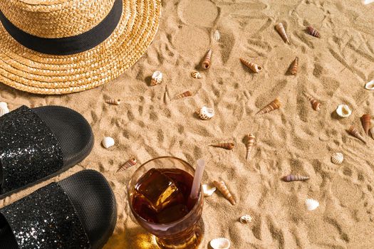 Summer beachwear, flip flops, hat, cold drink in a glass and seashells on sand beach. Top view. Copy space. Still life mockup flat lay